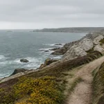 Sentier côtier balisé sur une pointe de granit bretonne, ajoncs en fleurs et mer gris-vert sous un ciel couvert